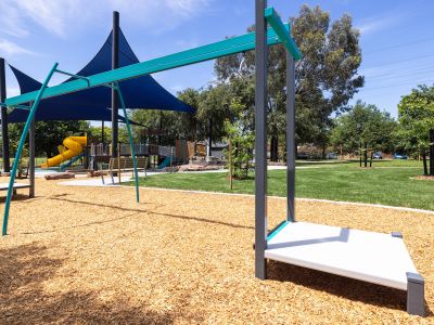 A photo of Howard Dawson Reserve's upgraded playground showing swings, shade sails, and a yellow slide on blue rubber and mulch surface