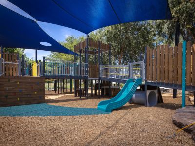 A photo of Howard Dawson Reserve's upgraded playground showing swings, shade sails, and a yellow slide on blue rubber and mulch surface